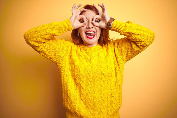 Mulher ruiva bonita vestindo camisola de inverno em pé sobre fundo amarelo isolado fazendo gesto ok como binóculos furando a língua para fora, olhos olhando através dos dedos. Expressão louca
. - Foto, Imagem
