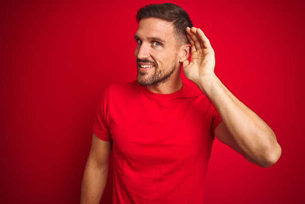 Mladý pohledný muž v neformálním tričku na červeném izolovaném pozadí se usmívá s rukou přes ucho a poslouchá drby nebo drby. Koncept hluchoty. - Fotografie, Obrázek