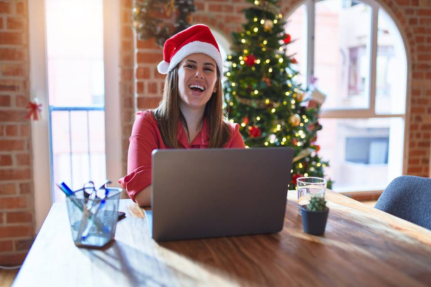 Красива жінка сидить за столом, працюючи з ноутбуком у капелюсі Санта-Клауса на різдвяному щасливому обличчі, посміхаючись схрещеними руками, дивлячись на камеру. Позитивна особа
. - Фото, зображення