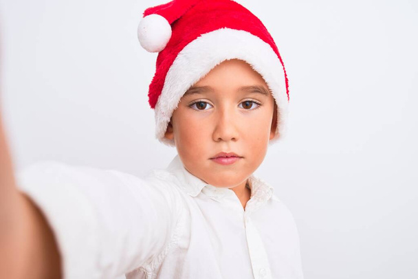 Hermoso niño con sombrero de Navidad Santa hacer selfie sobre fondo blanco aislado con una expresión de confianza en la cara inteligente pensando en serio
 - Foto, Imagen