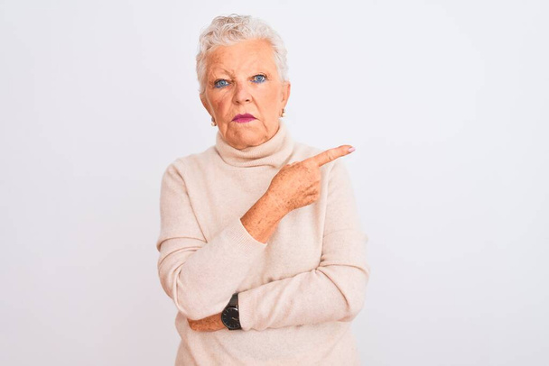 Starší šedovlasá žena v rolákovém svetru, stojící nad izolovaným bílým pozadím, ukazující ukazováčkem na stranu s reklamou, vážnou a klidnou tváří - Fotografie, Obrázek