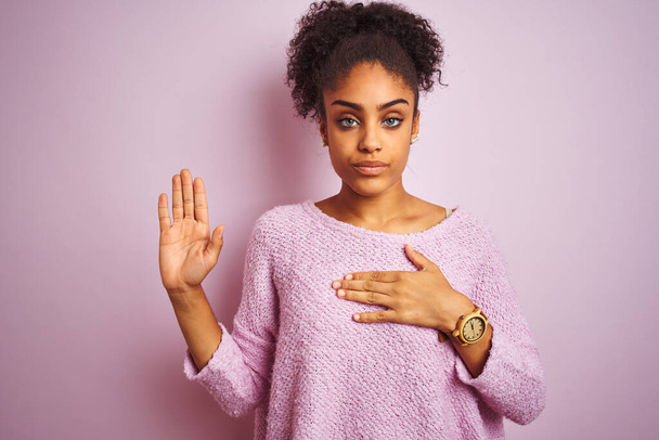 Młoda Afroamerykanka w zimowym swetrze stojąca nad odizolowanym różowym tłem przysięgająca ręką na klatkę piersiową i otwartą dłoń, składając przysięgę lojalności. - Zdjęcie, obraz