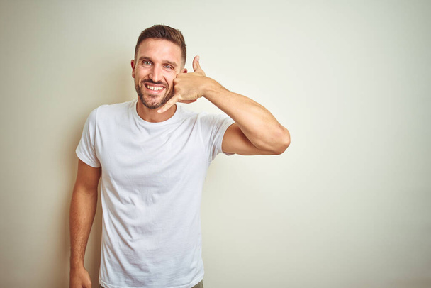 Молодой красивый мужчина в обычной белой футболке на изолированном фоне улыбается, делая телефонный жест рукой и пальцами, как будто разговаривает по телефону. Коммуникационные концепции
. - Фото, изображение