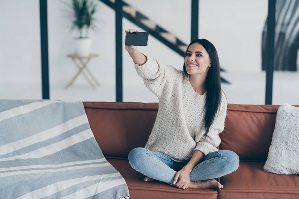 Portret van positief vrolijk meisje hold mobiele telefoon selfie foto zitten op divan in huis modern appartement feel content dragen gebreide trui denim jeans - Foto, afbeelding