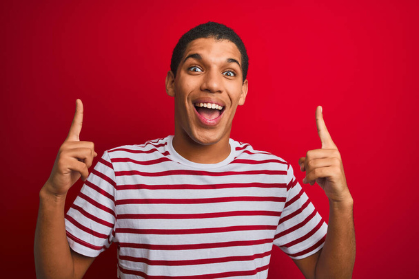 Jeune homme arabe beau portant t-shirt rayé sur fond rouge isolé souriant étonné et surpris et pointant vers le haut avec les doigts et les bras levés
. - Photo, image