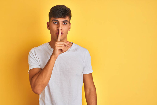 Young indian man wearing white t-shirt standing over isolated yellow background asking to be quiet with finger on lips. Silence and secret concept. - Photo, Image