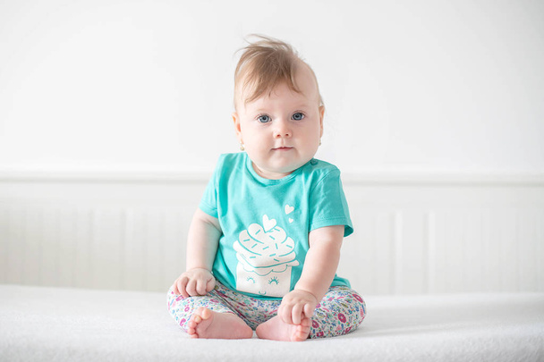 Portret uroczej białej dziewczynki z niebieskimi oczami, spoglądającej spokojnie w kamerę, z ciekawością, interaktywnością lub dociekliwością i siedzącej w nieskazitelnym białym łóżeczku niemowlęcym trzymając palce u stóp - Zdjęcie, obraz