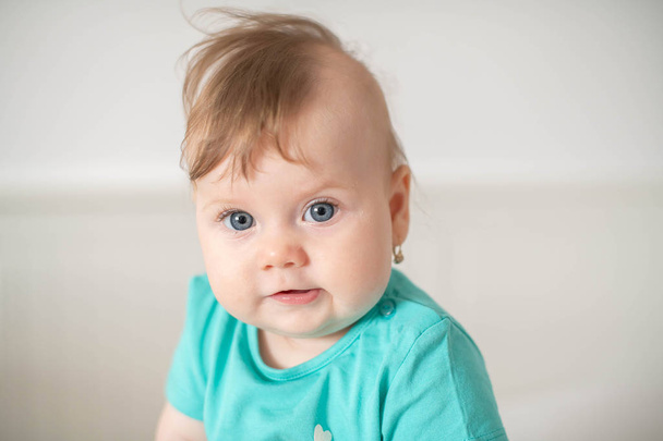 Portrait of adorable Caucasian baby girl with blue eyes, looking at the camera calmly, with curiosity, interactivity or inquisitiveness and sitting in a white baby cot; cute baby expressions concepts - Photo, Image