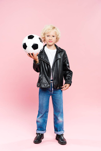 kind in lederen jas houden van voetbal en kijken naar de camera op roze achtergrond  - Foto, afbeelding