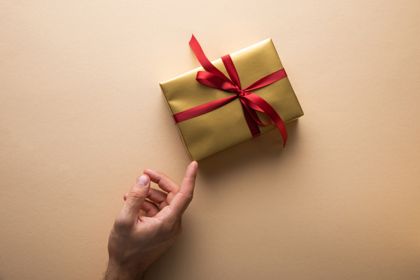 cropped view of man touching golden gift box with red ribbon on beige background - Photo, Image