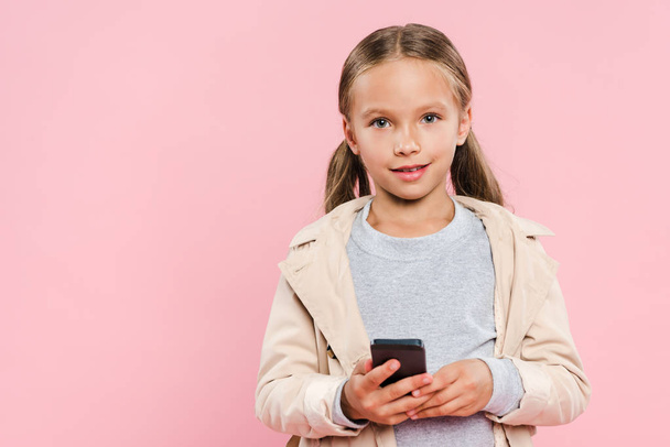 χαμογελαστό παιδί σε φθινοπωρινή στολή κρατώντας smartphone απομονωμένο σε ροζ - Φωτογραφία, εικόνα