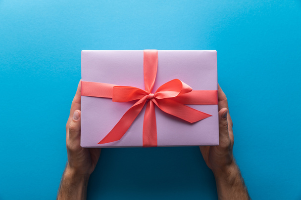 cropped view of man holding violet gift box with red ribbon on blue background - Foto, afbeelding