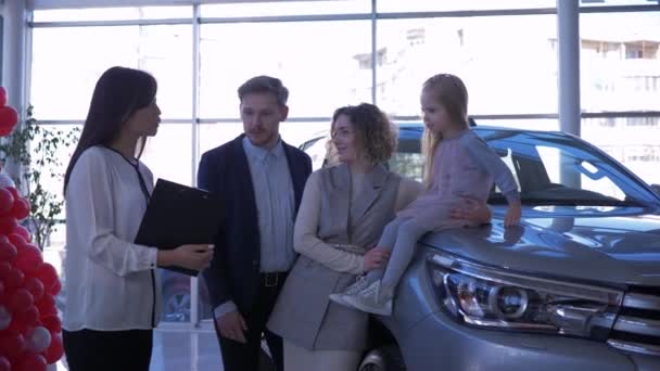 Автосалон, сім'я молодих покупців з дитиною спілкується з менеджером автомобіля про купівлю нового автомобіля в центрі продажу автомашин - Кадри, відео