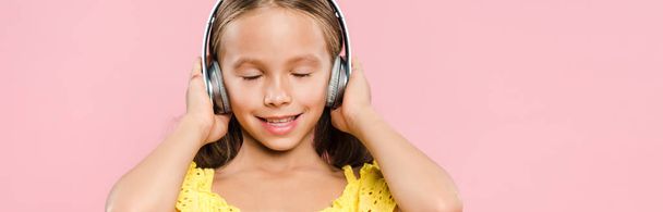 panoramic shot of smiling kid with headphones listening music isolated on pink - Photo, Image