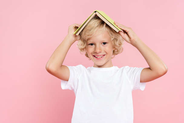 smiling kid holding book and looking at camera isolated on pink - Fotoğraf, Görsel