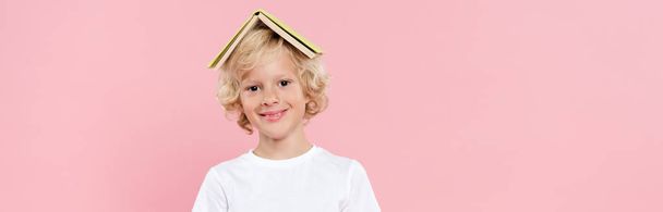 panoramic shot of smiling kid with book on head isolated on pink - Фото, изображение