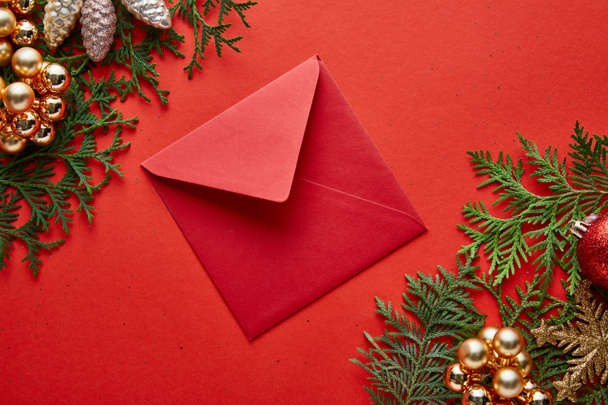 top view of red envelope near shiny Christmas decoration on green thuja branches on red background - Photo, image