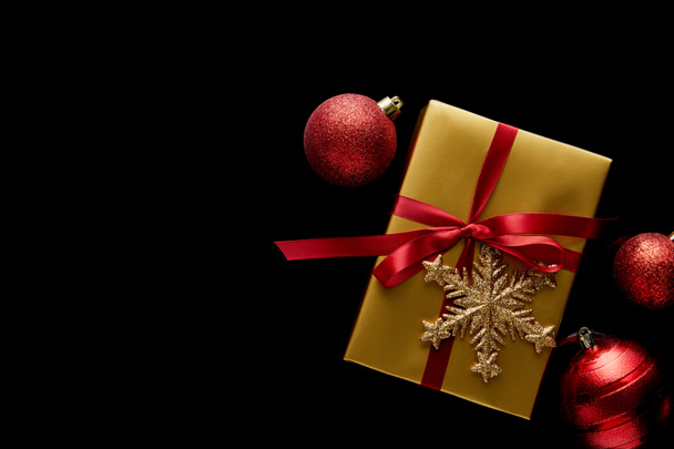 felső nézet fényes arany karácsonyi ajándék piros szalaggal és hópelyhek közelében baubles elszigetelt fekete - Fotó, kép