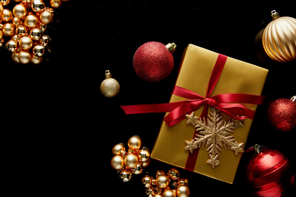 top view of shiny golden Christmas gift with red ribbon and snowflake near baubles isolated on black - Foto, Imagem