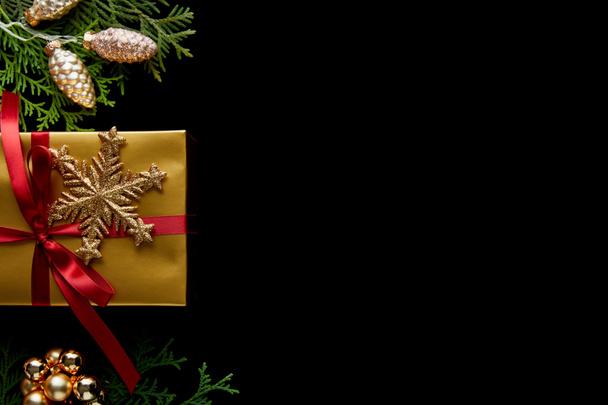 vrchní pohled na lesklé zlaté vánoční dekorace, zelené větve thuja a dárek izolované na černé s kopírovacím prostorem - Fotografie, Obrázek