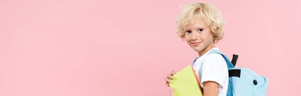 panoráma lövés mosolygós iskolás fiú hátizsák holding könyv elszigetelt rózsaszín - Fotó, kép