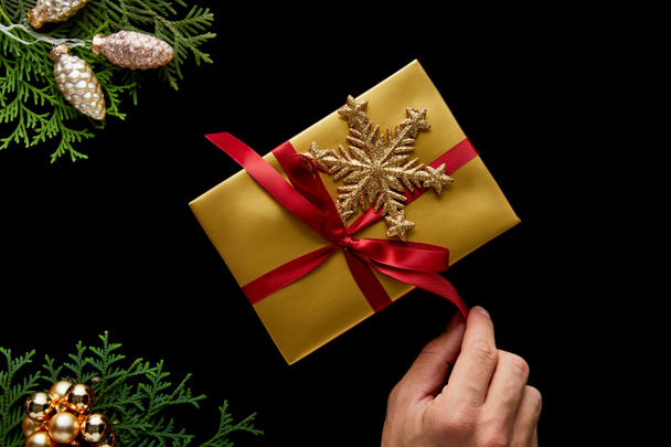 обрезанный вид человека развязывающий лук на подарок рядом с блестящим золотым украшением Рождества, зеленые ветви туи изолированы на черном
 - Фото, изображение