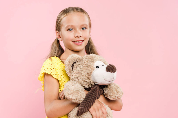 glimlachend kind kijken naar camera en houden teddybeer geïsoleerd op roze - Foto, afbeelding