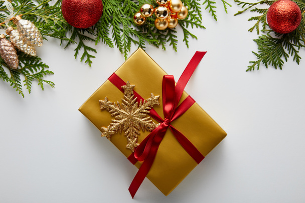 Yeşil thuja dalları üzerindeki parlak altın ve kırmızı noel süslemesinin üst görüntüsü, beyaz üzerinde kartaneleri olan paketlenmiş hediye. - Fotoğraf, Görsel