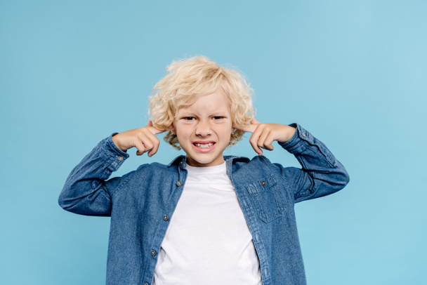 irritated and cute kid looking at camera and touching ears isolated on blue  - Foto, Imagem