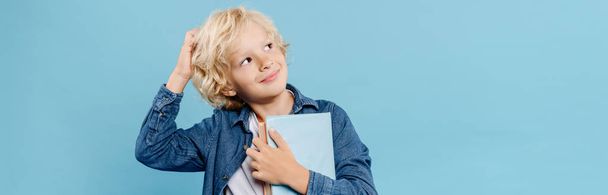 panoramic shot of smiling and dreamy kid holding books isolated on blue  - Foto, afbeelding