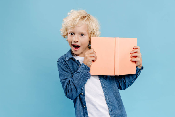 shocked and cute kid holding book and looking at camera isolated on blue  - Photo, Image
