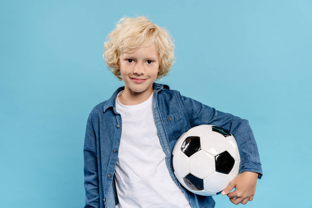 smiling and cute kid looking at camera and holding football isolated on blue  - Zdjęcie, obraz