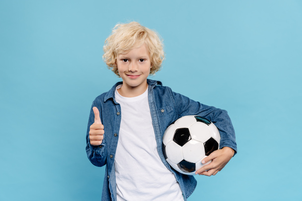 smiling and cute kid showing like and holding football isolated on blue  - Foto, immagini