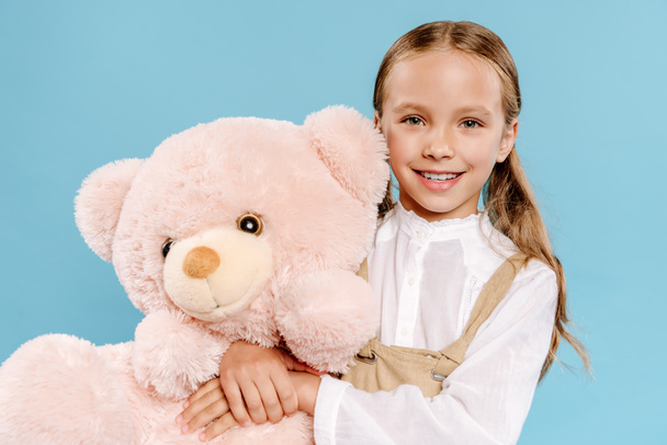 smiling and cute kid looking at camera and holding teddy bear isolated on blue  - Photo, image