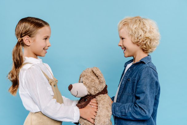 side view of smiling and cute kids holding teddy bear isolated on blue  - Foto, imagen