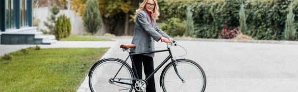 tiro panorâmico de atraente empresária sorrindo e segurando bicicleta
  - Foto, Imagem