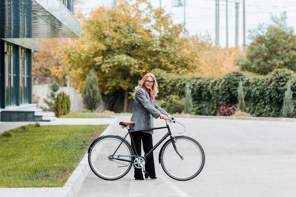 attractive businesswoman in coat and glasses smiling and holding bike  - Foto, immagini