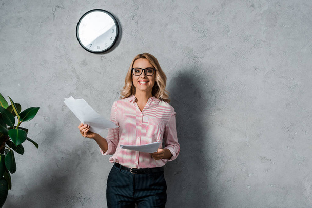 attractive businesswoman in shirt and glasses smiling and holding papers  - Foto, Imagem