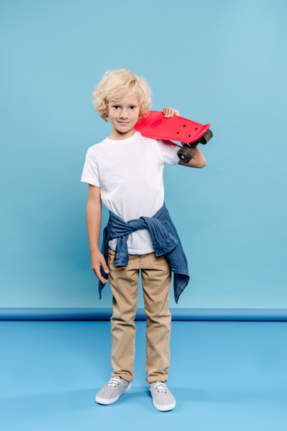 smiling and cute kid looking at camera and holding penny board on blue background  - Foto, Imagen