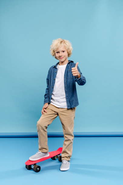 smiling and cute kid showing like and standing on penny board on blue background  - Фото, зображення