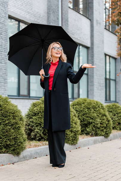 attractive businesswoman in coat and glasses holding black umbrella with outstretched hand  - Фото, зображення