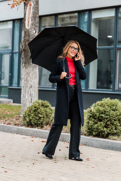 attractive businesswoman in coat and glasses holding black umbrella and talking on smartphone  - Fotoğraf, Görsel