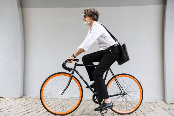 Photo of modern old businessman riding bicycle over gray wall - Zdjęcie, obraz