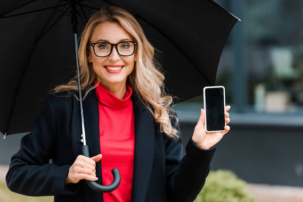 attraktive und lächelnde Geschäftsfrau im Mantel mit Regenschirm und Smartphone  - Foto, Bild