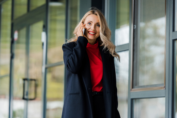 atraktivní a usmívající se podnikatelka v černém kabátě mluví na smartphone  - Fotografie, Obrázek