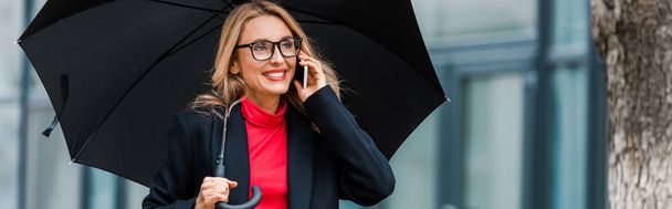 plano panorámico de atractiva y sonriente empresaria en abrigo negro sosteniendo paraguas y hablando en smartphone
  - Foto, imagen