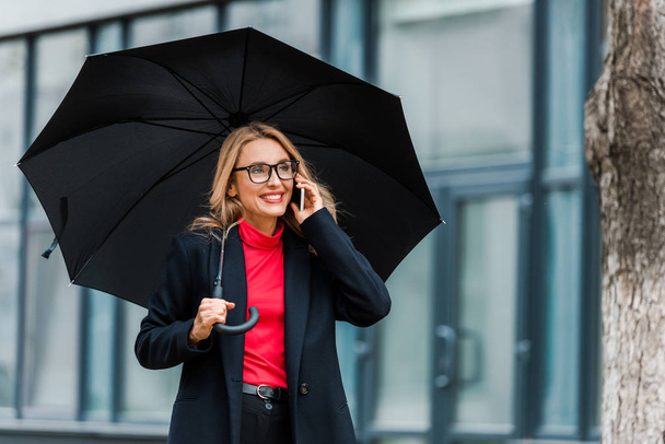 привлекательная и улыбающаяся деловая женщина в черном пальто держит зонтик и разговаривает по смартфону
  - Фото, изображение