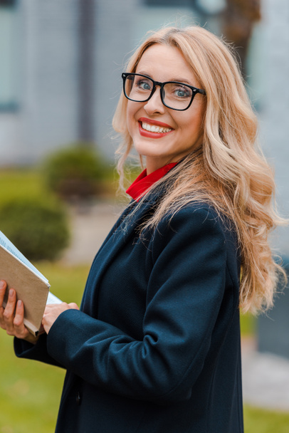 attraktive und lächelnde Geschäftsfrau in schwarzem Mantel und Brille mit Buch  - Foto, Bild