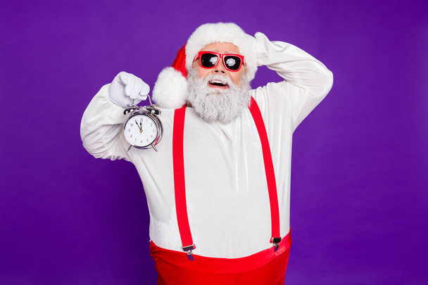 Photo of funny overweight santa father holding alarm clock remind its newyear oclock overjoyed wear sun specs hat suspenders isolated purple background - Фото, зображення
