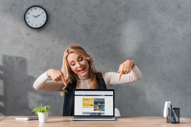 KYIV, UKRAINE - AUGUST 16, 2019: smiling businesswoman pointing with fingers at laptop with booking website  - Fotografie, Obrázek